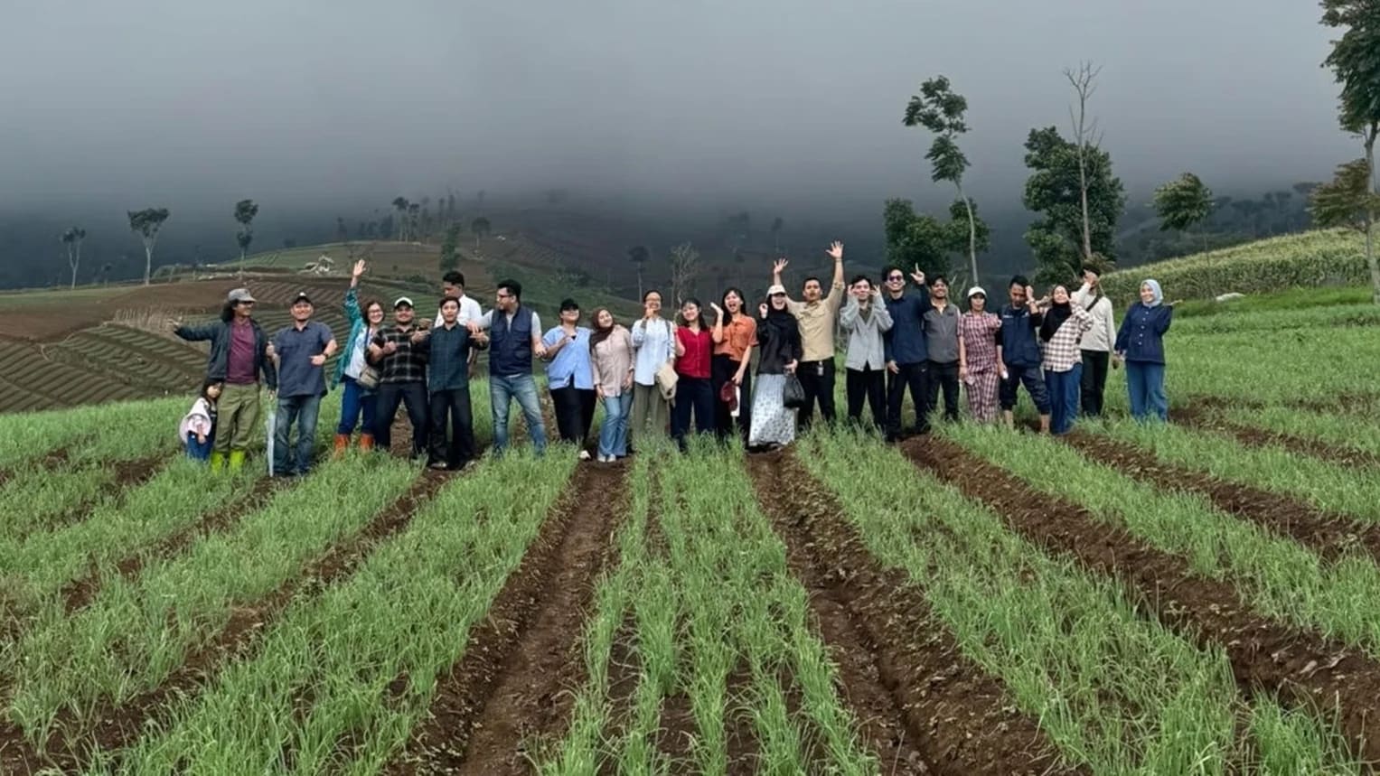 Sekelompok sekitar dua puluh orang tersenyum dan melambaikan tangan sambil berdiri bersama di lahan tanaman muda berwarna hijau di lereng bukit, dengan kabut menyelimuti perbukitan di belakangnya.