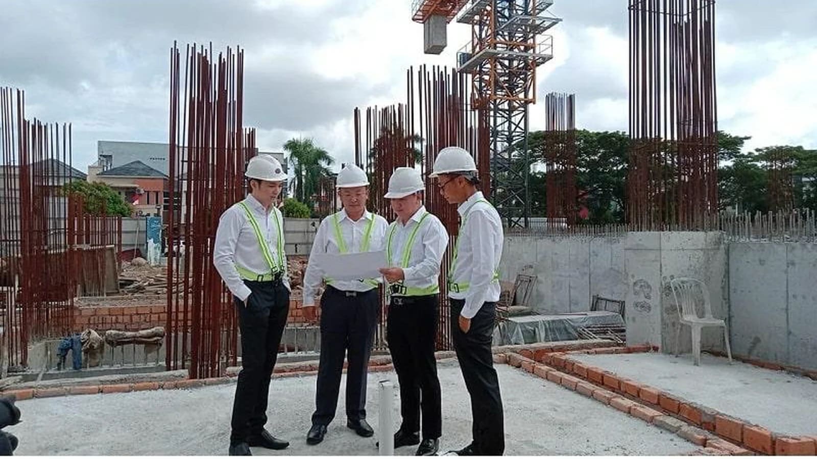 Empat orang mengenakan helm proyek putih dan rompi reflektif sedang meninjau dokumen bersama di lokasi konstruksi dengan besi tulangan yang terbuka dan sebuah menara crane.