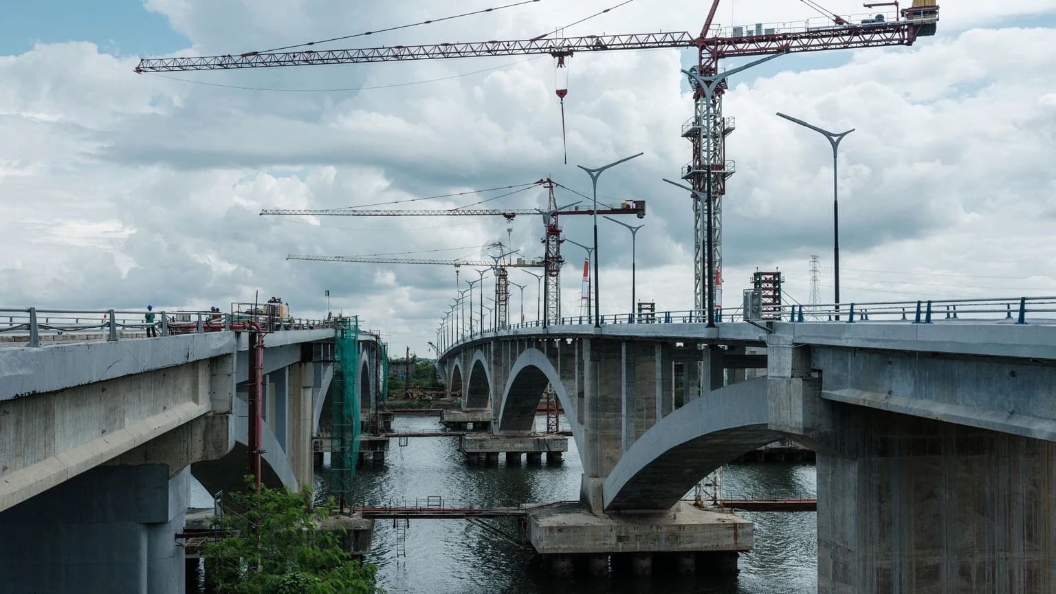 Dua jembatan lengkung beton yang sedang dibangun di atas sungai, dengan derek menara, lampu jalan, dan perancah di bawah langit berawan; beberapa pekerja berhelm keselamatan berdiri di dek sebelah kiri.