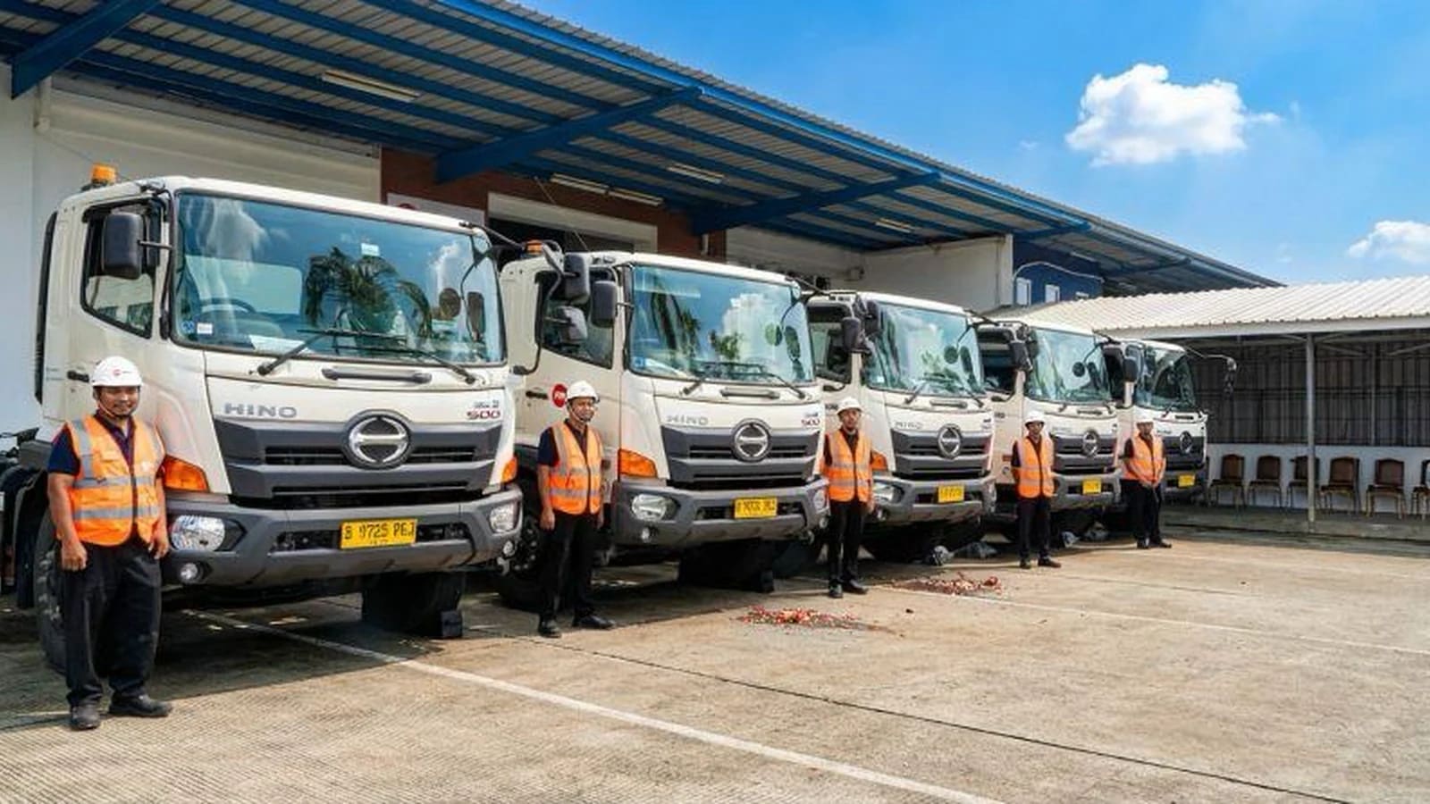 Deretan truk besar berwarna putih terparkir di depan gudang beratap baja biru, dengan seorang pekerja berompi keselamatan oranye dan berhelm putih berdiri di samping setiap truk.