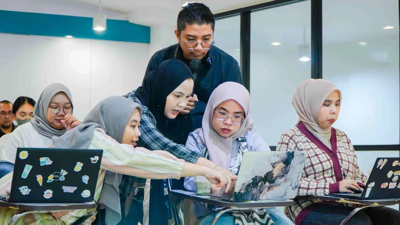 Sejumlah profesional muda bekerja dengan laptop di ruang pelatihan; seorang perempuan mencondongkan badan untuk menunjuk layar rekannya, sementara seorang pria yang memegang mikrofon memperhatikan dari belakang.