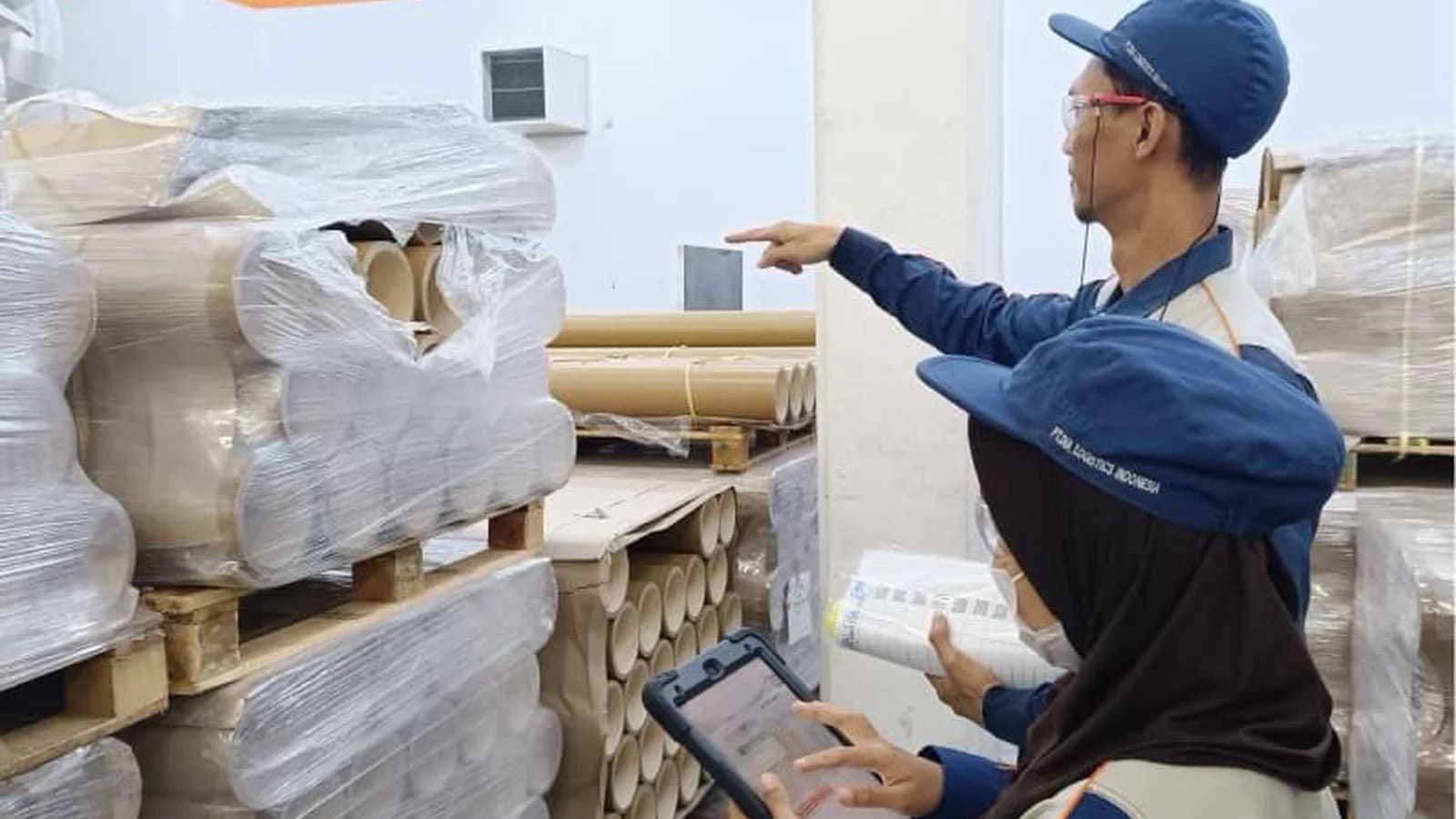 Dua pekerja bertopi dan berseragam biru memeriksa tumpukan tabung karton terbungkus plastik di atas palet dalam sebuah gudang; satu orang menunjuk, sementara yang lain memegang tablet dan dokumen.
