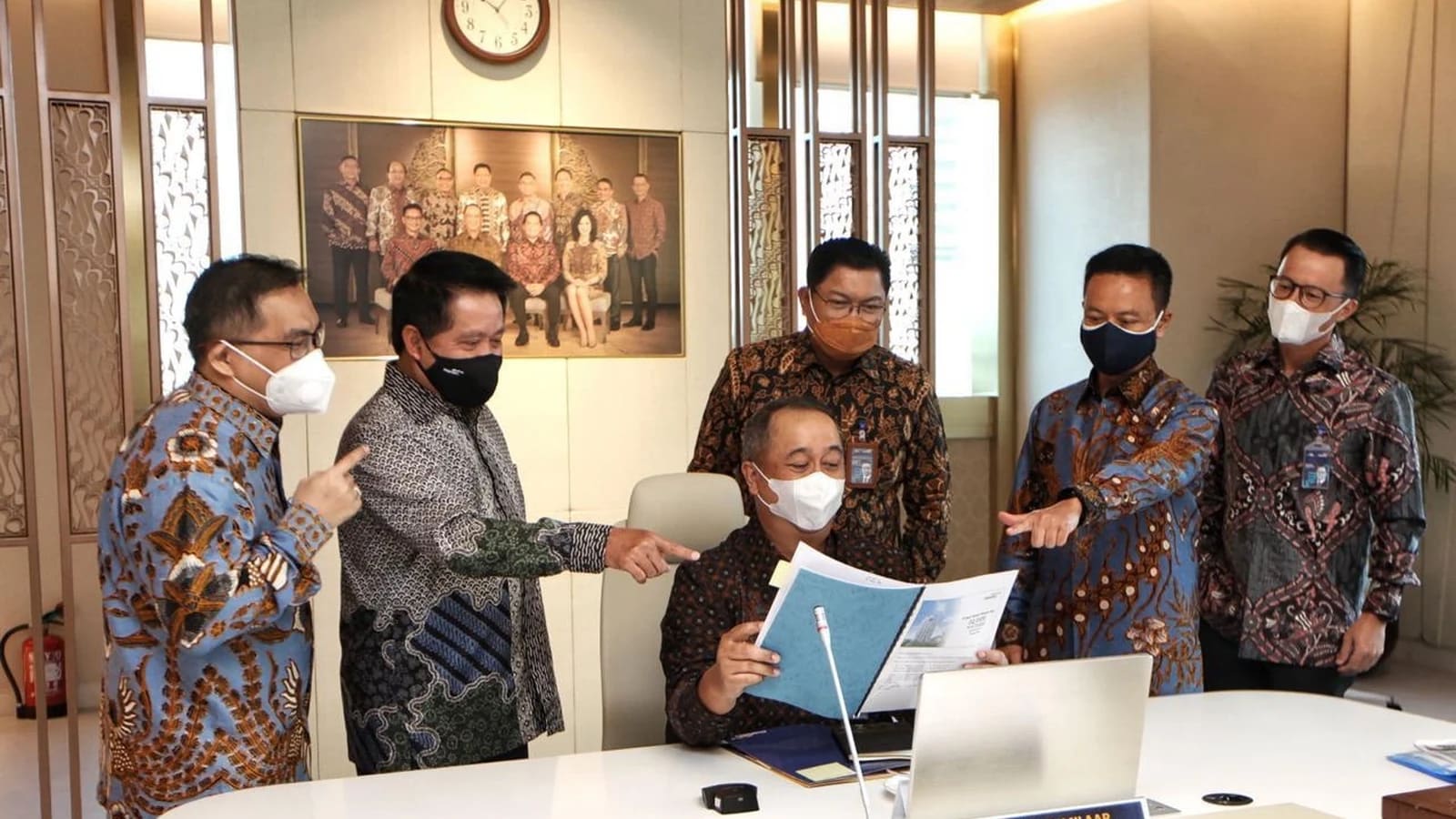 Lima pria berkemeja batik dan bermasker berkumpul di sekitar seorang rekan yang duduk sambil membaca dokumen di meja kantor, dengan foto bersama berbingkai dan jam dinding di belakang mereka.