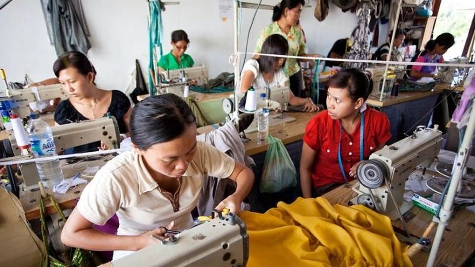Para perempuan bekerja dengan mesin jahit di sebuah bengkel garmen kecil, dengan kain kuning cerah di latar depan dan gulungan benang di atas meja kerja.